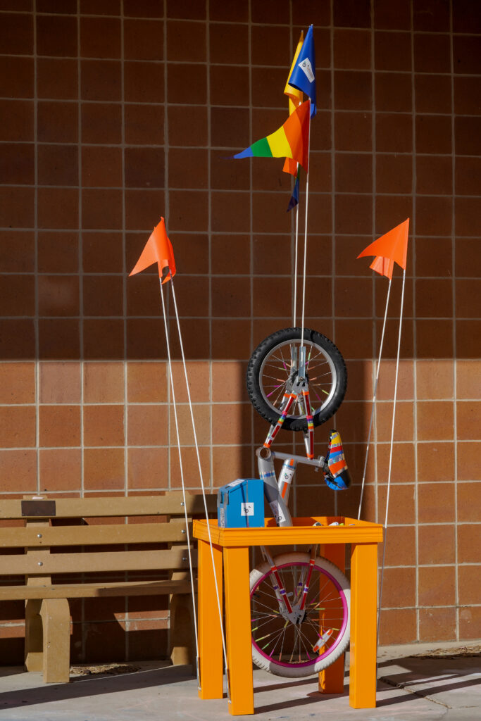 Image of orange bike sculptural table featuring bicycle parts, wheels, and multi-colored flags.
