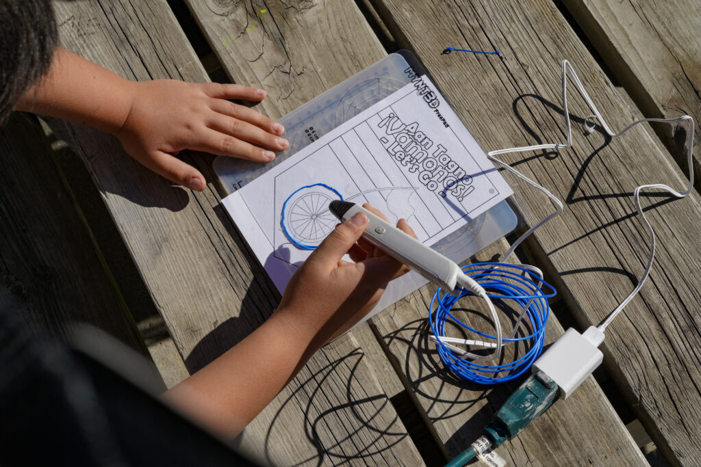 Image of the hands of a young person using a 3D printer pen to outline a phrase on a piece of paper that reads “Aan Tagno, Vamonos, Let’s Go.”