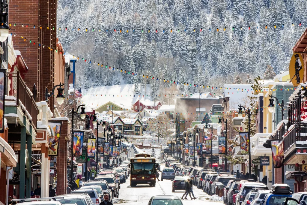 Free trolley bus on a busy street filled with lots of cars parked. Holiday lights, trees, cute colorful houses, and a mountain in the background Park City, Utah