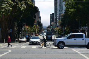 A cyclist and pedestrian in a long crosswalk are dwarfed by a Ford F-150 pick-up traveling in the same direction