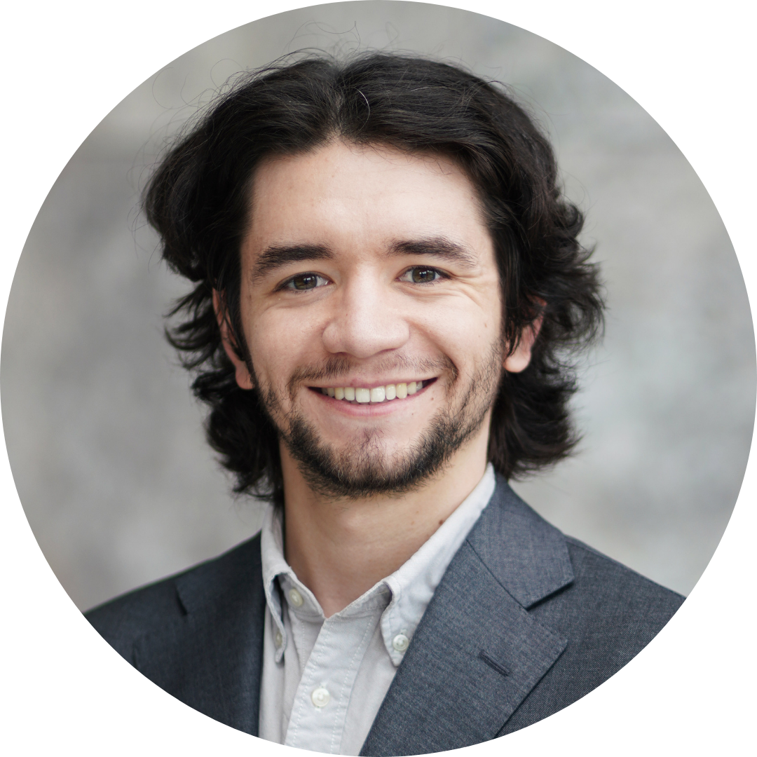 Headshot of Corrigan Salerno, wearing a gray suit jacket and navy tie against a brick and industrial background outside.